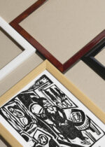 Widok z góry na cztery drewniane ramki, w jednej z nich oprawiony plakat czarno-biały Kanoniczka przy stole do szycia 1912 Kirchner